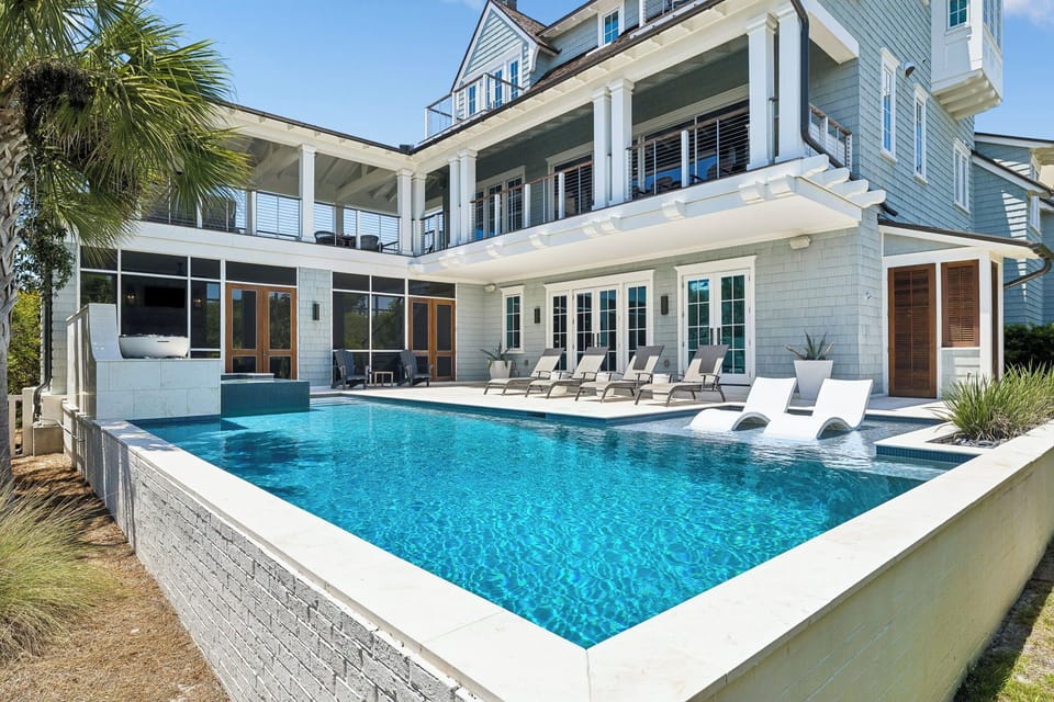 Elegant coastal property featuring an elevated pool deck with lounge seating, multiple balconies, and tropical landscaping under clear blue skies.