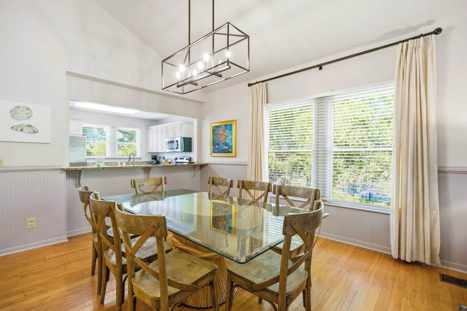 Natural wood grains and a glass-topped table catch the morning glow in this airy dining space. High ceilings and a modern chandelier create an open, breezy atmosphere for shared meals near the sun-drenched windows.