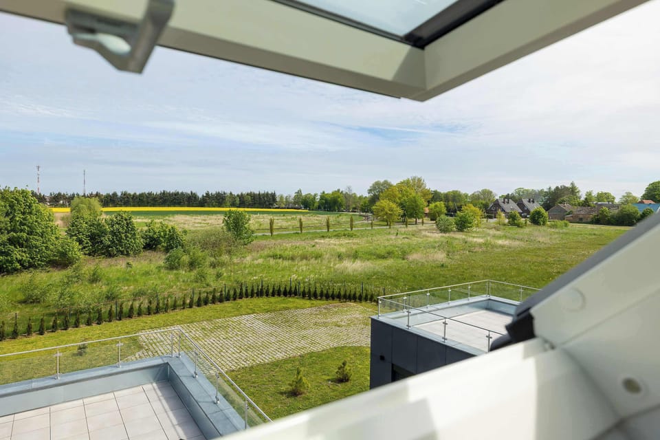 A view looking out through an open skylight window reveals a sprawling green field under a vast blue sky. A glass balcony railing is visible in the foreground, overlooking the rural landscape.
