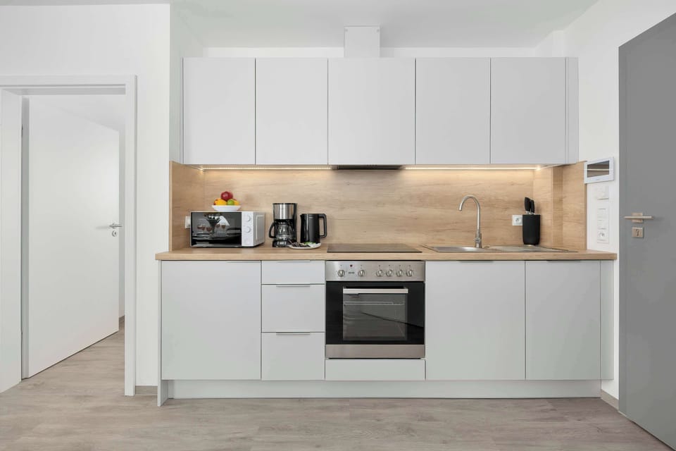 A clean, modern kitchen area showcases plain white cabinetry and a light wood backsplash. An integrated oven and stove are neatly fitted beneath a built-in extractor hood.