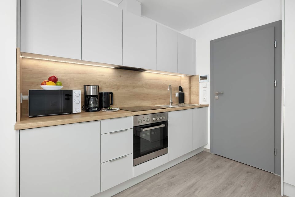 The modern kitchenette is shown with a black microwave and a coffee maker placed on the countertop. A grey door stands at the end of the counter line, completing the contemporary look.