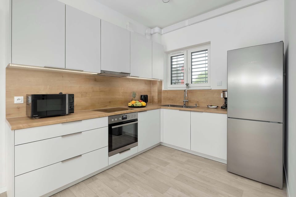 A modern kitchenette with white cabinets, wooden countertops, and stainless steel appliances. Large windows make the space bright and inviting.