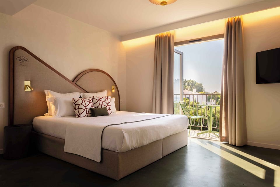 White-linened bed with curved headboard — the south-facing balcony with green chairs is through the glass door