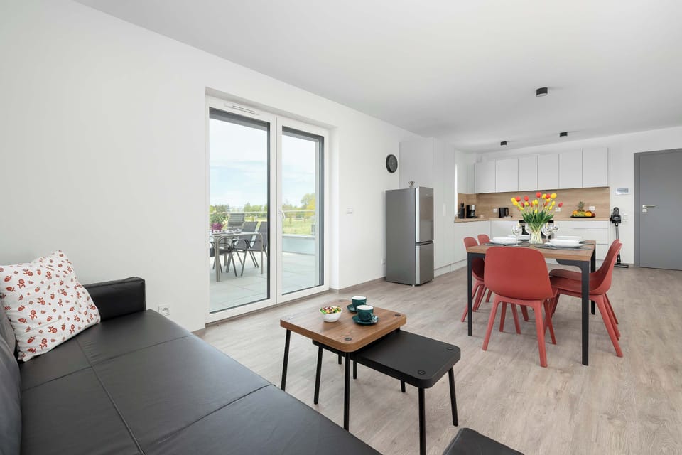An open-concept layout reveals a seamless flow between the living sofa area and a dining set with red chairs in the background. Glass doors offer a direct exit to the large outdoor terrace.