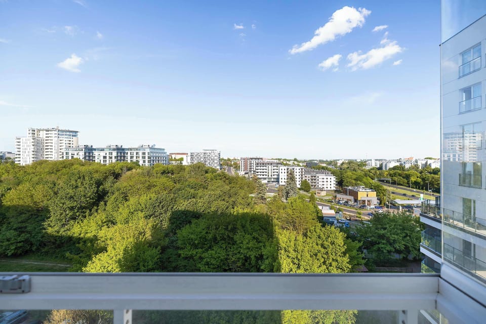 A wide balcony view overlooking treetops, residential buildings, and distant city structures under a blue sky. Glass railings provide an unobstructed panorama.