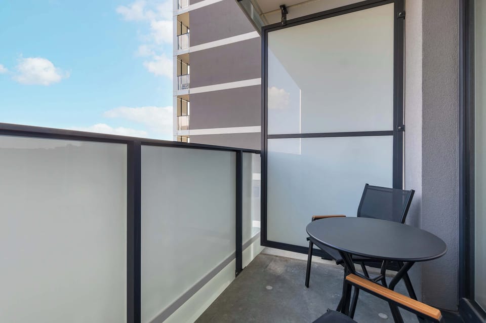 A small balcony with seating space for outdoor relaxation. The view includes nearby residential buildings in a quiet neighborhood.