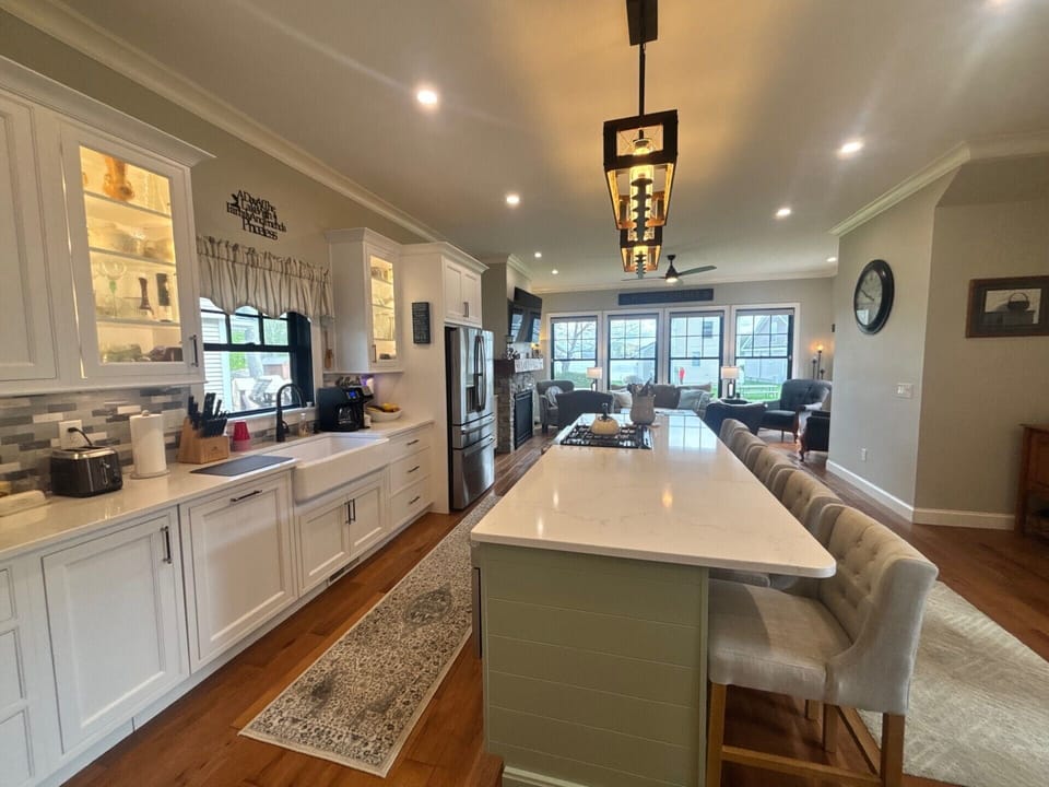 Gather around the spacious kitchen island where morning coffee and evening meals unfold naturally. White cabinetry, gleaming counters, and warm wood floors flow seamlessly into the inviting living space beyond.