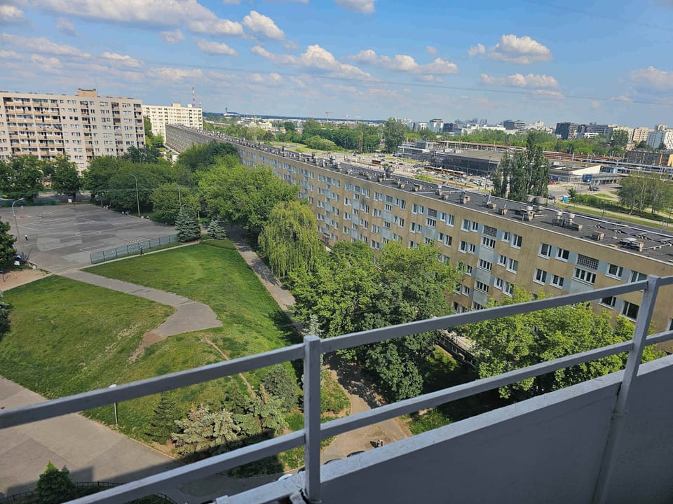 An impressive panoramic view from the balcony overlooks an expansive green park, a large outdoor parking lot, and distant city buildings. A clear blue sky with fluffy white clouds stretches across the horizon.