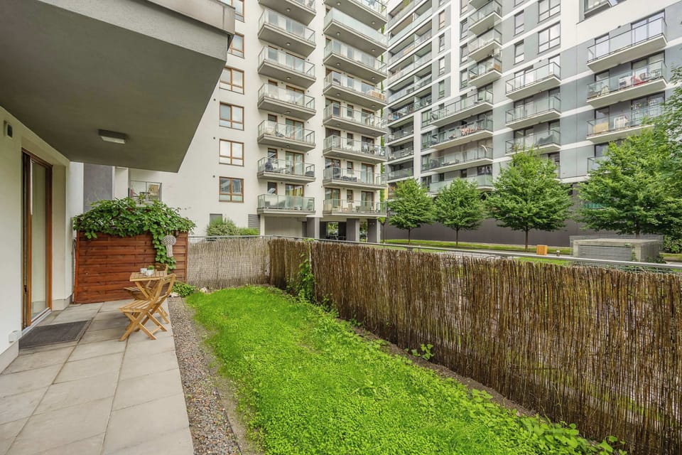 A peaceful private garden with a paved terrace, outdoor seating, and neatly maintained greenery surrounded by modern apartment buildings. The enclosed outdoor area offers a quiet spot for relaxing in the fresh air.