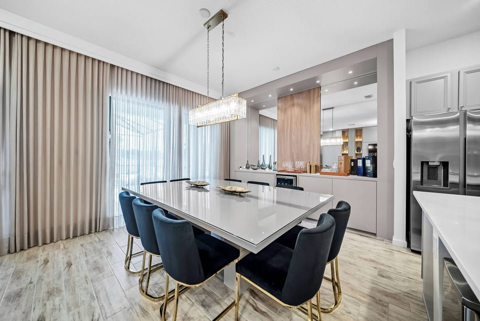Welcome to this stylish kitchen with a chic white marble table and plush navy chairs, perfect for dining. Large windows with soft curtains fill the space with natural light, beautifully highlighting the warm wood cabinetry.