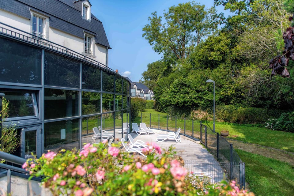 White sun loungers line the poolside terrace, framed by trees and the glass walls of the pool building.