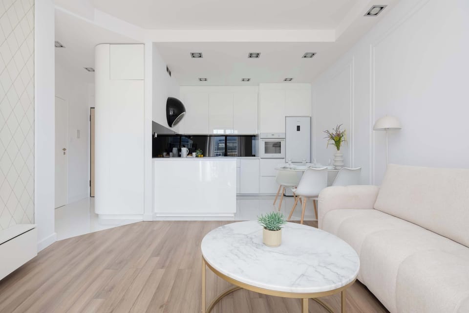 A bright, modern living room with an open-plan kitchen in the background, featuring white cabinetry and a clean, minimalist design. The space feels spacious, elegant, and filled with natural light.
