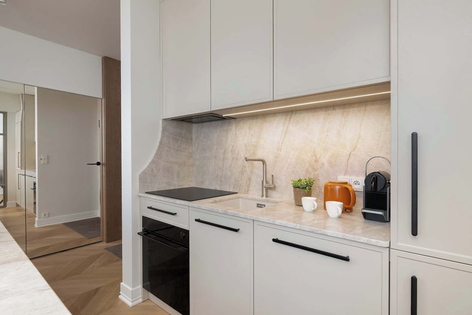 A sleek kitchenette features light-colored cabinetry, built-in appliances, and elegant stone-effect finishes. The minimalist design creates a clean and sophisticated look.
