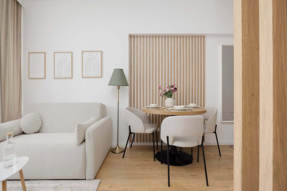 A cozy sitting space with neutral furnishings, framed wall art, and a small dining setup beside the window. Wooden accents complement the light-colored décor.
