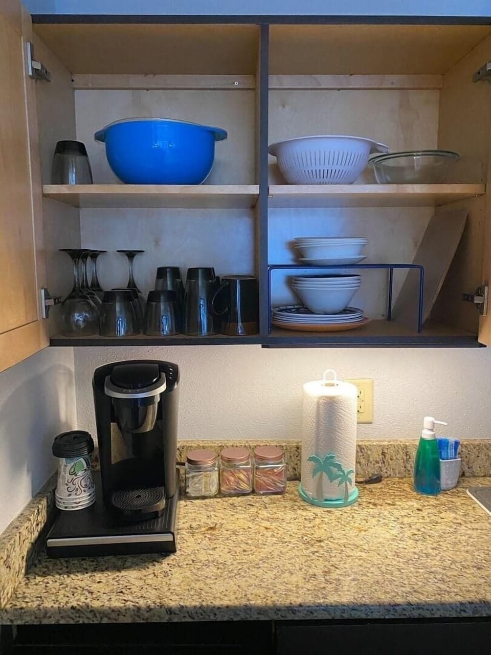 Well-stocked kitchen cabinet open above a granite countertop, displaying assorted dishes, bowls, and glassware alongside a coffee maker and paper towel roll.