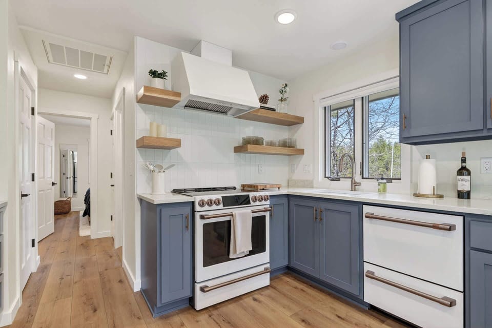 Your fully-equipped kitchen features sleek blue-gray cabinetry, modern appliances, natural light from a garden-view window, and warm wood flooring—perfect for preparing home-cooked meals during your stay.
