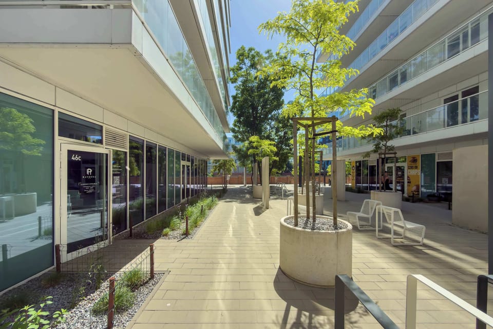 A wide pedestrian area between contemporary apartment buildings featuring outdoor seating and landscaped elements. Sunlight highlights the clean urban design.