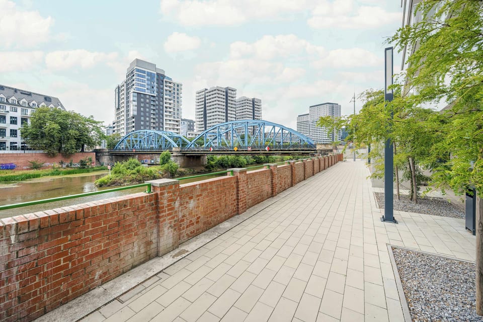 A riverside pedestrian walkway bordered by brick walls and lined with trees and modern buildings. Open views and greenery create a pleasant urban setting.