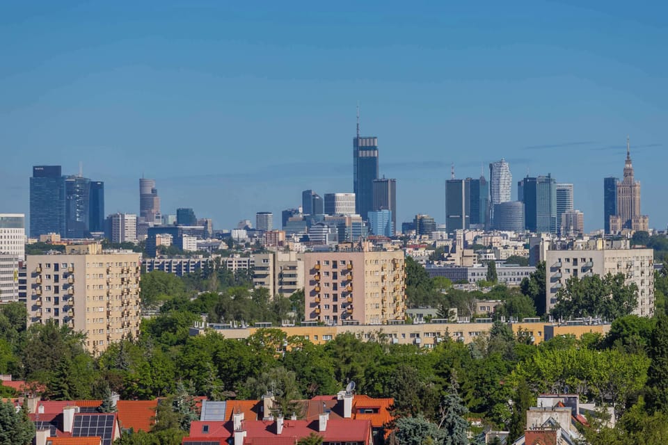 A panoramic city view with modern skyscrapers visible in the distance. Green areas and residential buildings can be seen in the foreground.