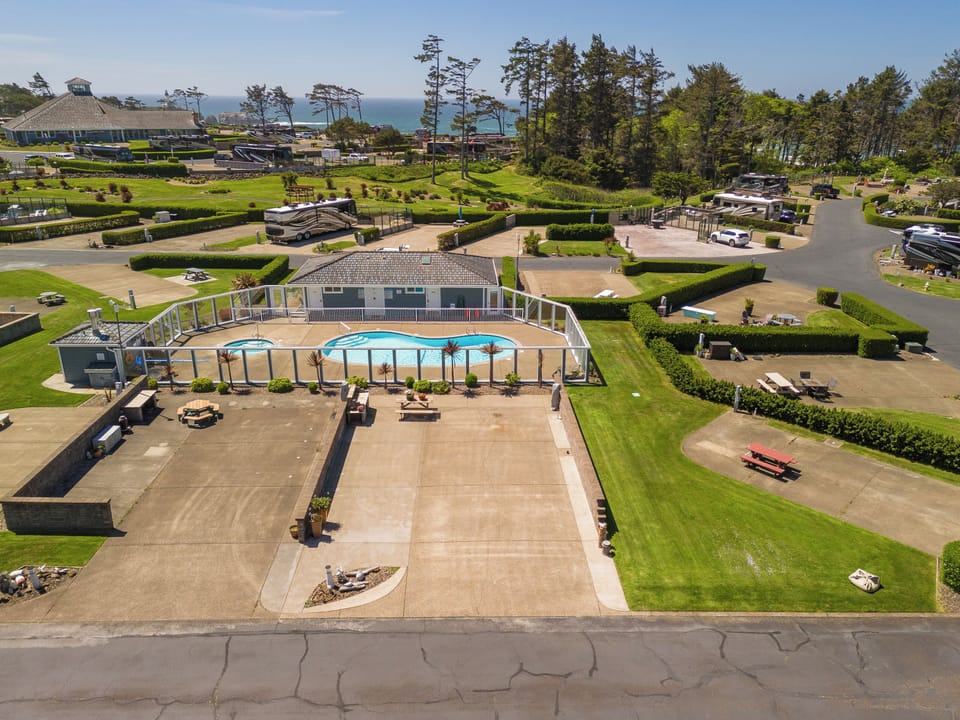 Aerial view reveals a resort property featuring an enclosed pool area surrounded by manicured lawns, mature pines, and sea views beyond. The landscaped grounds include parking areas and recreational spaces within a coastal setting.