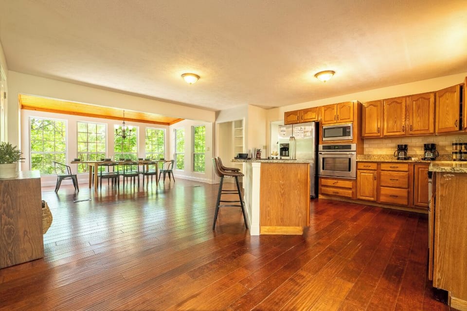 Your spacious kitchen flows into a sunlit dining area with forest views, where gleaming wood floors and warm wood cabinetry create a welcoming space for gathering and meals.
