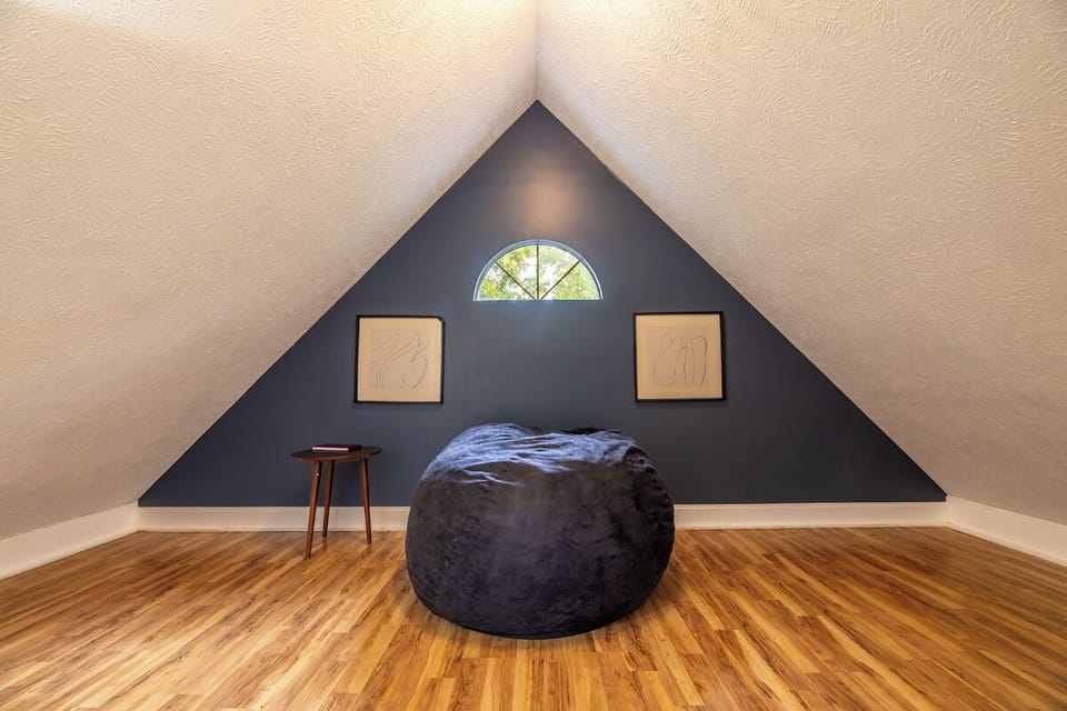 Unwind in your private retreat, where soft natural light filters through the arched window, highlighting the cozy bean bag chair and warm wood floors beneath the dramatic vaulted ceiling.