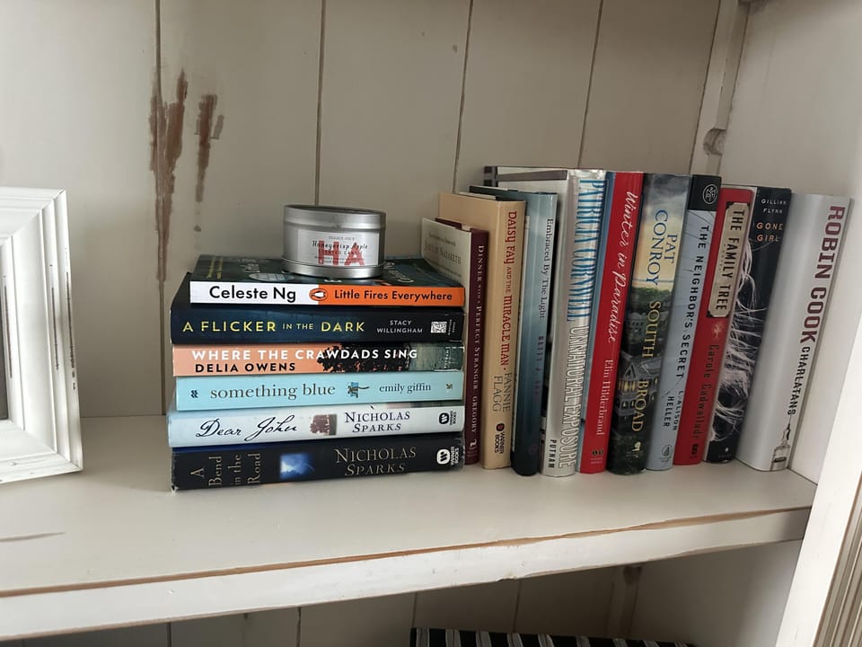 Bookshelf with a variety of novels and a small scented candle on a white wooden shelf with visible wall stains.