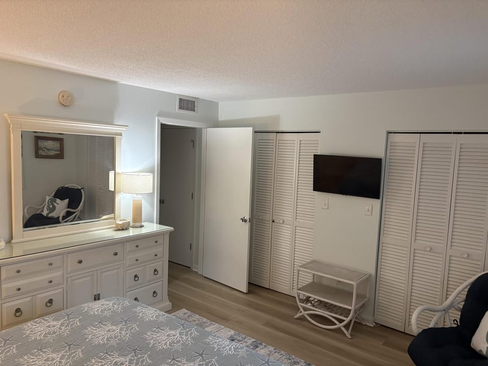 Bedroom features white louvered closets, a wall-mounted TV, a white dresser with mirror, and light wood flooring.