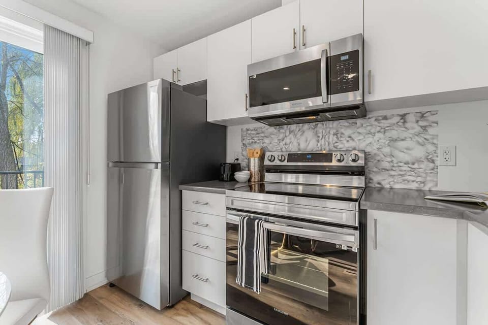 Sleek kitchen with stainless steel appliances, modern cabinetry, polished marble backsplash, built-in microwave, stylish oven, elegant utensils, and natural light streaming through large glass doors.