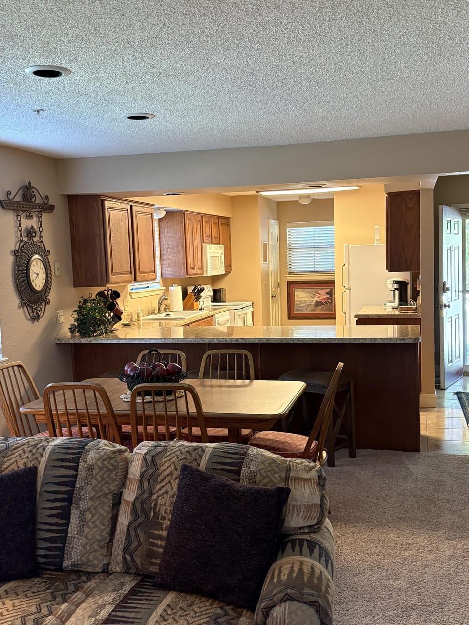 Wood dining table with six chairs sits beside a granite countertop kitchen featuring oak cabinets and white appliances.