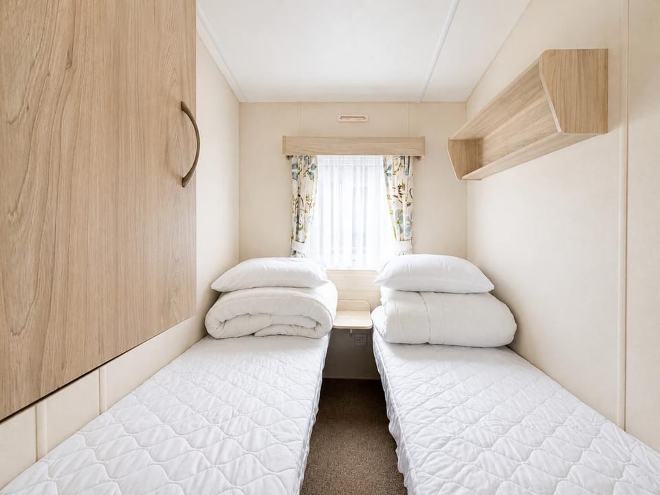 Twin beds with white quilts face a window framed by floral curtains, flanked by light wood shelves and a wardrobe.