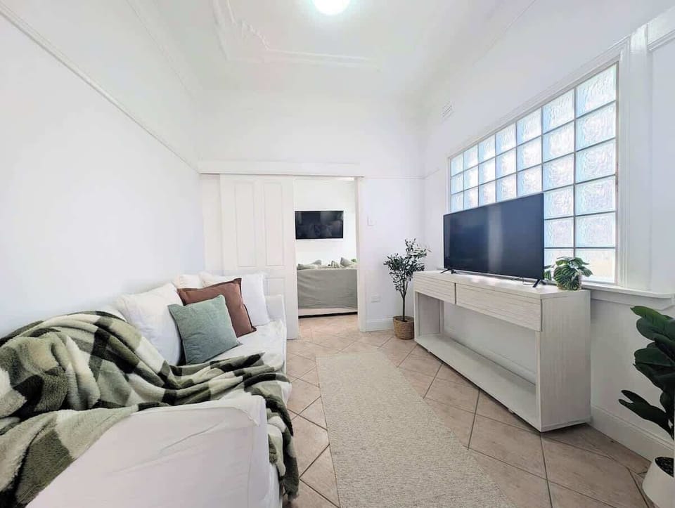 Relax in this bright living space, featuring a cozy sofa styled with colorful pillows and a plaid blanket, a flat-screen TV on a long white console, a glass block window wall, and lush potted greenery.