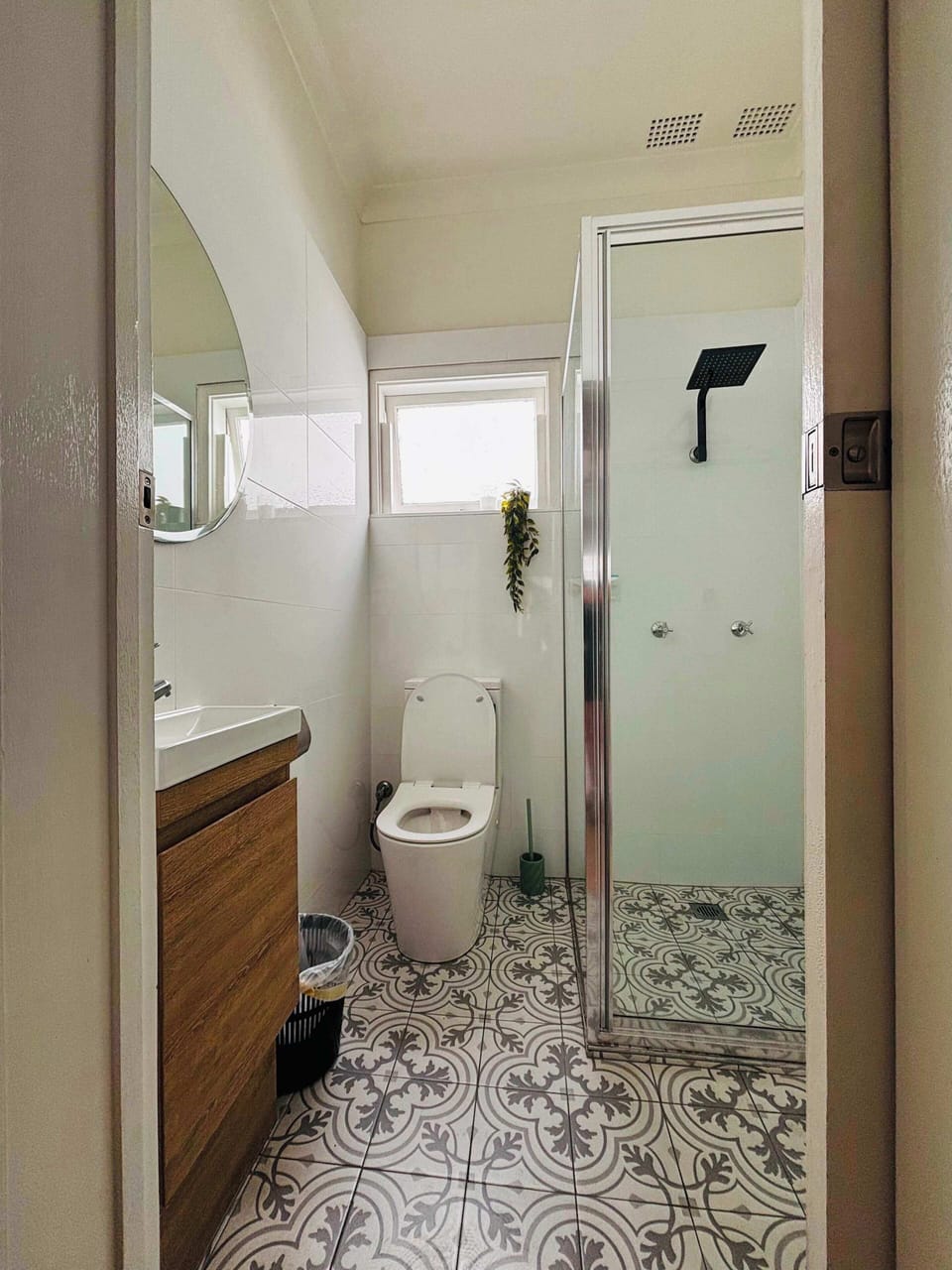Refresh in this well-appointed bathroom, featuring eye-catching patterned floor tiles, a wood-grain vanity, a modern toilet, and a glass-enclosed walk-in shower with a black rain showerhead.