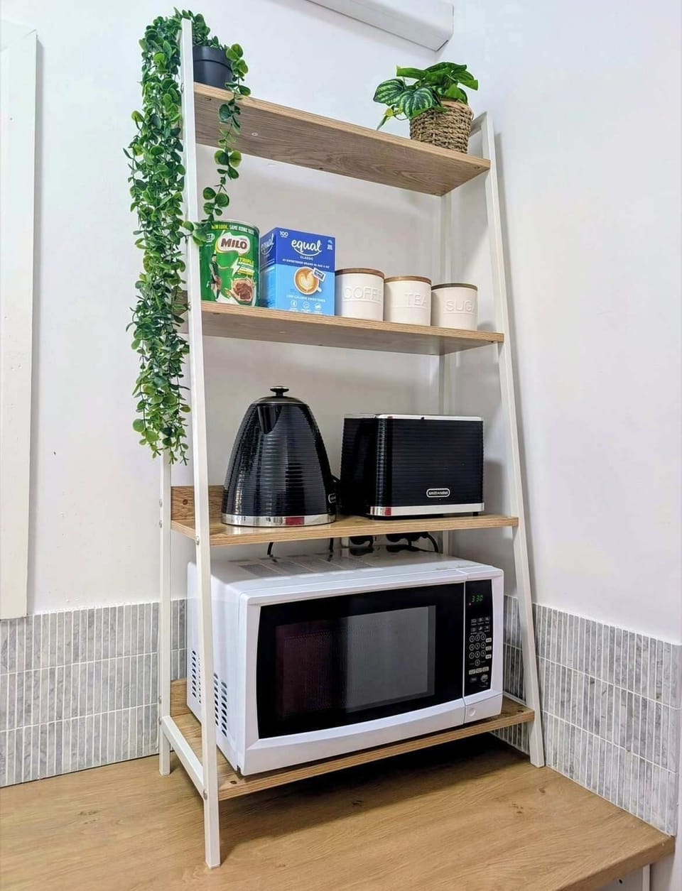 Organize with style on this modern multi-tier shelving unit, thoughtfully arranged with a microwave, electric kettle, small appliances, pantry essentials, and cascading greenery for a functional, space-saving kitchen setup.