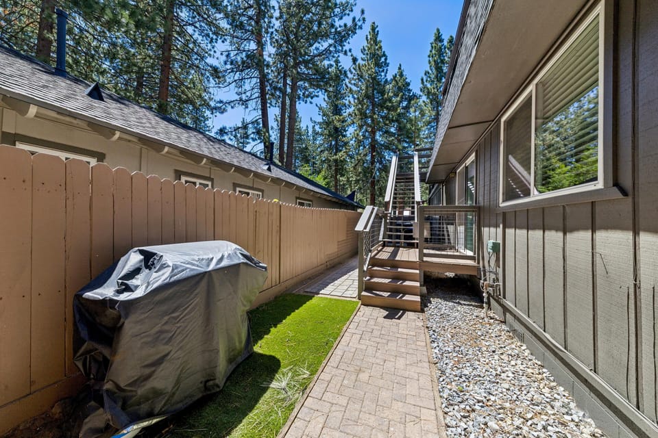 Side patio with brick pavers, artificial turf patch, covered grill, and metal staircase leading to upper deck.