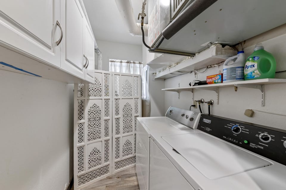Laundry room with white Maytag washer and dryer, open shelving, white cabinetry, and decorative folding screen.