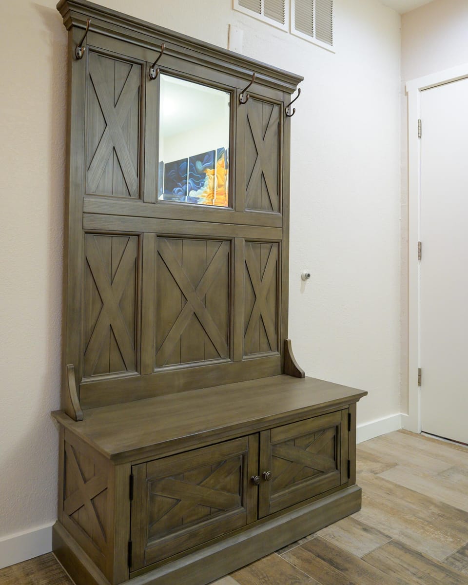 Entryway wooden hall tree with X-pattern panels, metal hooks, and a built-in storage bench on wood-look tile flooring.