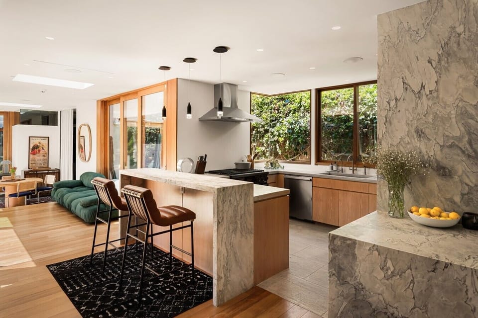 The kitchen flows seamlessly into the dining and living areas, with marble counters, warm wood tones, and natural light making the whole space feel connected.