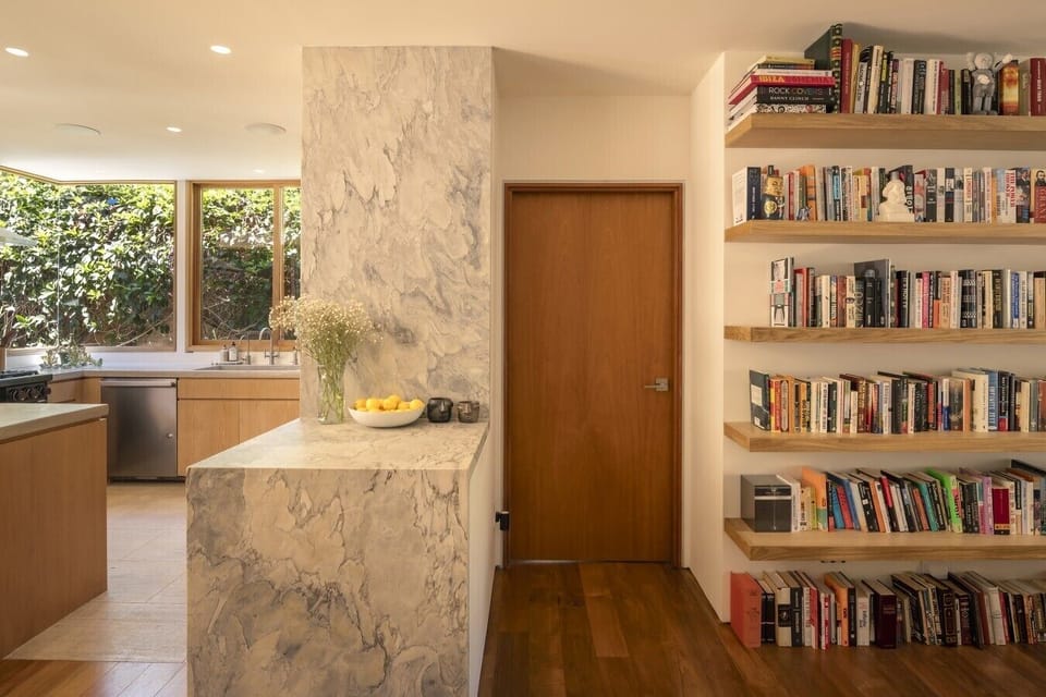 A well-equipped kitchen with marble countertops, garden window views, and a built-in bookshelf — functional, beautiful, and full of character.