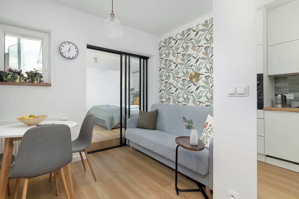 A dining corner sits beside patterned wallpaper and large glass doors leading outside. Soft gray seating complements the modern design.