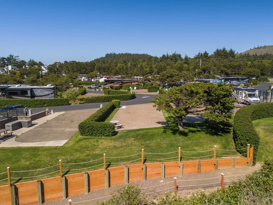 An RV park nestled in a scenic coastal area, surrounded by lush forests and rolling hills, featuring manicured lawns and landscaped grounds under clear blue skies.