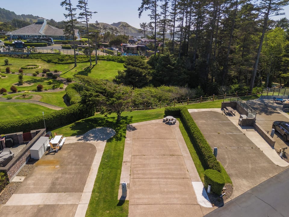 Aerial view of the property's spacious grounds featuring manicured lawns, mature trees, paved parking areas, and rolling coastal hills in the distance.