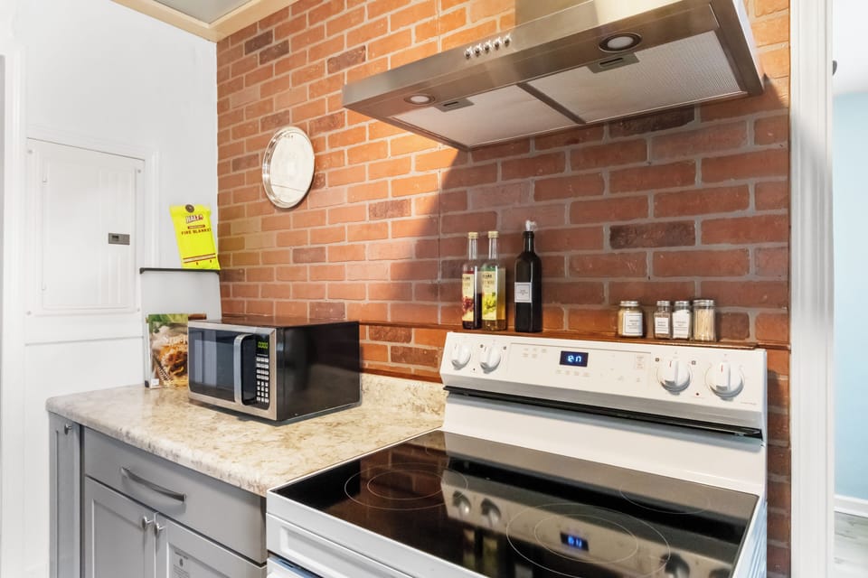 A kitchen cooking area featuring a stainless steel range hood mounted against a rustic exposed brick wall, with condiment bottles on the counter and a fire blanket nearby for safety.