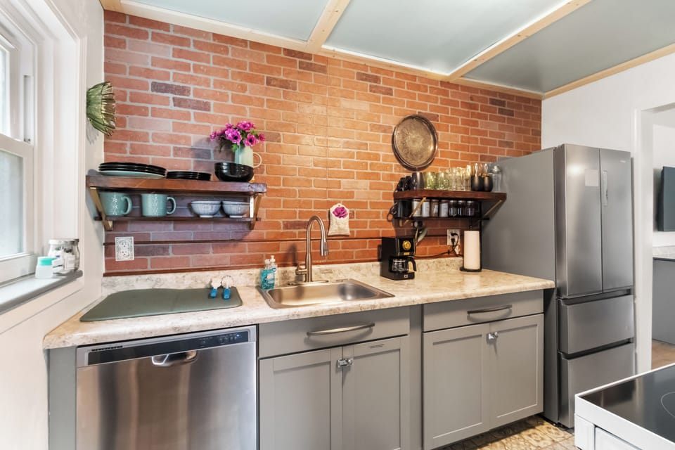 A charming kitchen with open wooden shelves display teal mugs, dishes, and a fresh flower arrangement, while a stainless steel sink, dishwasher, and fridge complete the functional yet stylish space.
