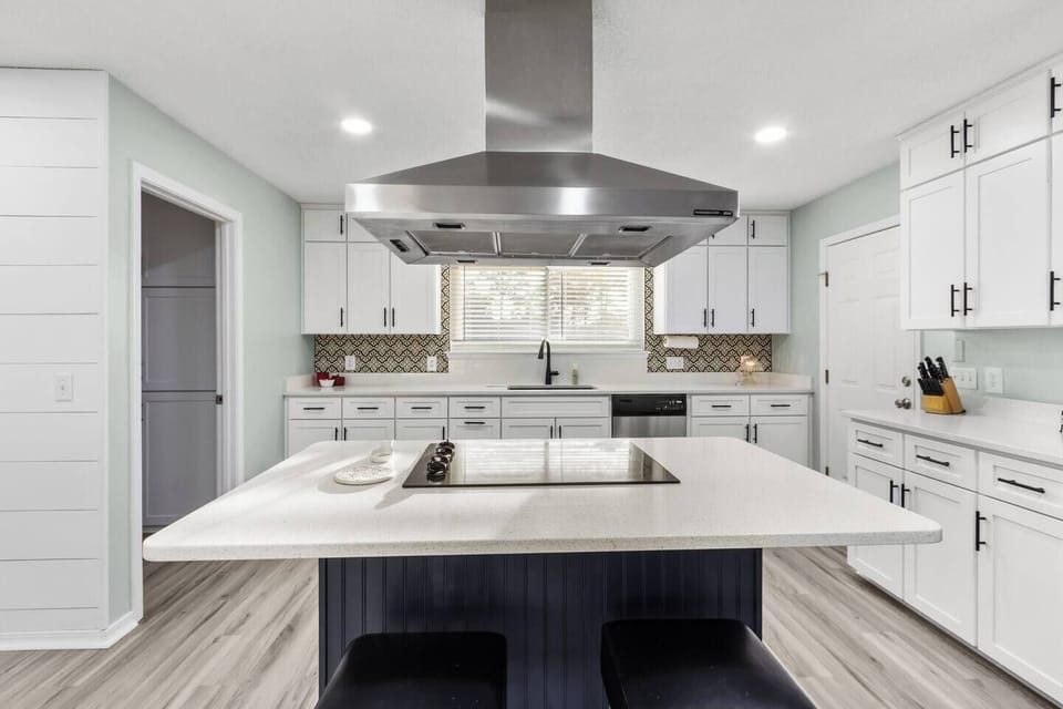 Modern kitchen with a large navy island, white quartz countertops, crisp white shaker cabinets, mosaic tile backsplash, and a stainless steel island hood. Light hardwood floors and recessed lighting complete this sleek, contemporary coastal kitchen.