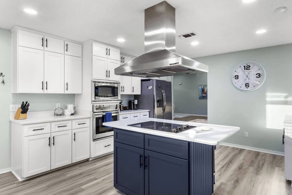 Navy island with cooktop, stainless steel hood, white shaker cabinets, and premium appliances including a double oven and French door refrigerator. Light hardwood floors complete this sleek coastal kitchen.