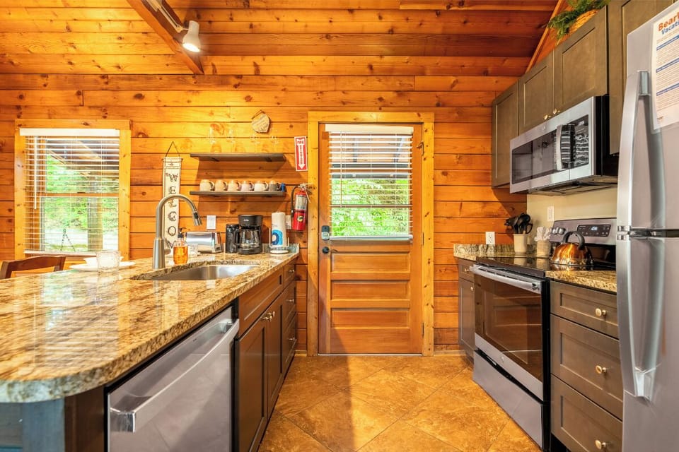 Your fully equipped kitchen blends rustic charm with modern convenience, featuring granite countertops, stainless steel appliances, and warm wood-paneled walls that create a welcoming space for preparing meals during your stay.