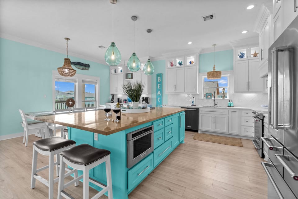 Bright kitchen with turquoise island, bar stools, white cabinets, stainless appliances, pendant lights and coastal decor.