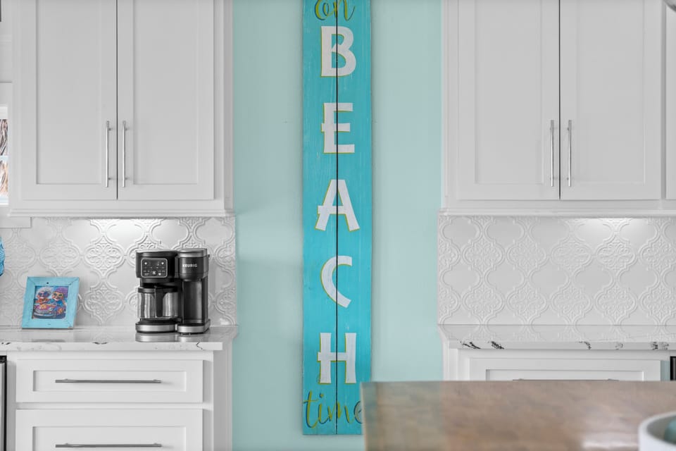 Coffee station with Keurig-style coffee maker, white cabinetry, decorative backsplash and beach-themed wall sign for easy morning coffee.