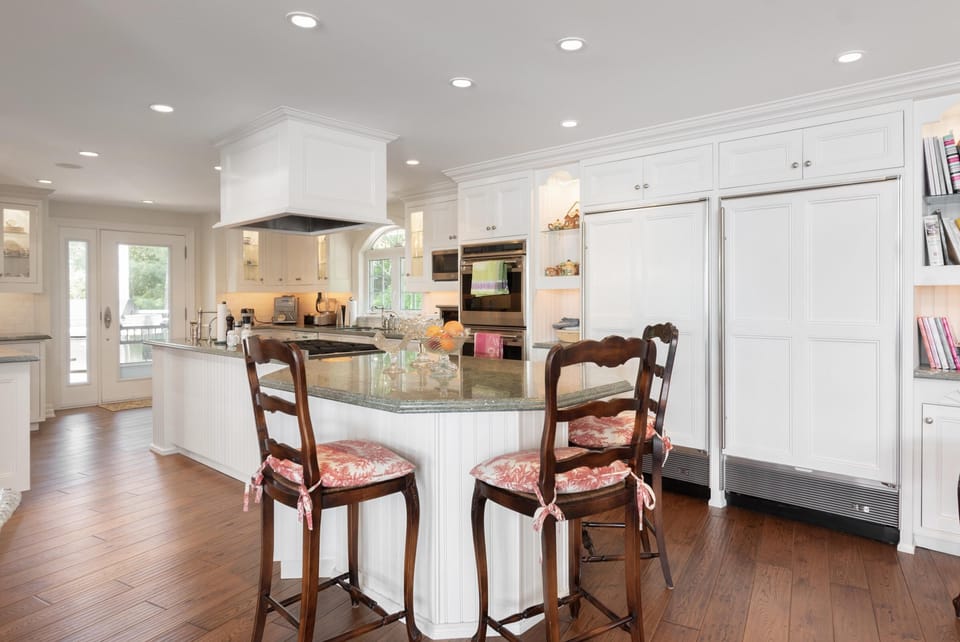 White cabinetry surrounds the granite-topped island with wooden stools tied with patterned cushions.